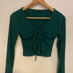 Grön ribbad croppad topp med knyt - Snygg grön croppad topp med lång ärm och ribbad struktur. Toppen har v-ringning och knyt framtill som ger en trendig look. Perfekt att matcha med höga jeans eller kjol.