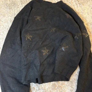Svart stickad tröja med stjärnor - Mysig svart stickad tröja med diskreta glittriga stjärnor över hela plagget. Tröjan har långa ärmar och bred ribbad mudd vid midjan och ärmsluten. Perfekt för dig som gillar en enkel men ändå lite extra look.