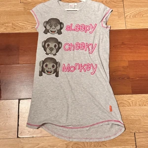 Grå t-shirtklänning med ap-emojis från h&m - Grå t-shirtklänning från Emoji med tryck av tre ap-emojis och texten 'Sleepy Cheeky Monkey' i rosa. Klänningen har korta ärmar med rosa kant och rund halsringning. Perfekt för en avslappnad stil.