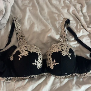 Svart spets-bh med broderade detaljer från Chantelle - Svart bh med vacker vit broderad spets längs kupor, axelband och nederkant. Bh:n har breda axelband och ger ett elegant och lyxigt intryck. Perfekt för dig som vill ha något extra fint under kläderna.