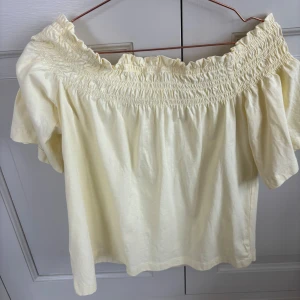 Gul offshoulder blus - Säljer en ljusgul offshoulder blus med smock upptill och korta ärmar. Toppen har en luftig och somrig känsla, perfekt för varma dagar. Den har en söt volangkant längs halsringningen och är tillverkad i ett mjukt bomullsliknande material.
