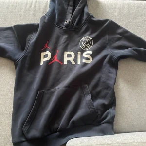 Svart Paris Saint-Germain x Jordan hoodie - Säljer en svart hoodie från Paris Saint-Germain x Jordan med stor logga på ryggen och tryck framtill. Tröjan har huva, magficka och ribbade muddar. Perfekt för dig som gillar sportig streetstyle och vill sticka ut med snygg design.