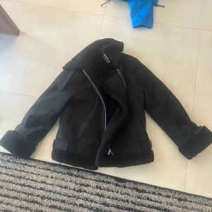 Svart jacka med fuskpälsfoder och bälte - Säljer en svart pufferjacka med mjukt fuskpälsfoder och bred krage. Jackan har dragkedja, stora spännen vid ärmsluten och bälte i midjan. Perfekt för kyliga dagar och riktigt snygg till jeans eller kjol.