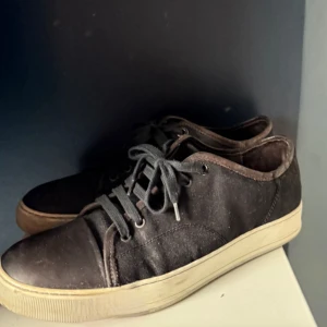 Svarta sneakers från Lanvin - Säljer ett par svarta sneakers från Lanvin med vit platt sula och snörning. Skorna har en klassisk och stilren design med rund tå och diskreta sömmar. Perfekta för dig som gillar minimalistisk stil.