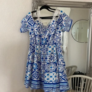 Blåvit kortklänning med spetsdetaljer - Säljer en blåvit kortklänning med unikt kakelinspirerat mönster och fina spetsdetaljer vid axelbanden. Klänningen har korta ärmar och en markerad midja som ger en feminin siluett. Perfekt för sommarens alla tillfällen!