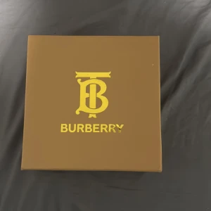 Burberry bälte med klassiskt rutmönster - Säljer ett Burberry bälte med det ikoniska beigea rutmönstret och svart läderdetalj. Bältet har ett guldfärgat spänne med Burberrys logotyp. Kommer i originalförpackning med dustbag och presentpåse.