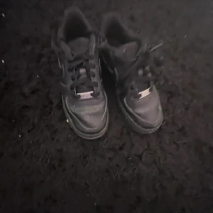 Svarta Nike Air Force 1 sneakers - Säljer ett par klassiska svarta Nike Air Force 1 sneakers med svart sula och svarta skosnören. Skorna har den ikoniska AF1-detaljen på snörningen och är tillverkade i skinn. Perfekta för dig som gillar streetwear och tidlös stil.