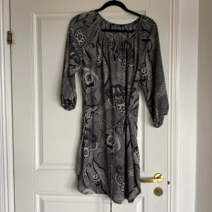 Mönstrad klänning från Gina Tricot - Säljer en snygg grå och svart mönstrad klänning från Gina Tricot. Klänningen har trekvartsärm, knytning vid halsen och ett paisleyinspirerat mönster. Perfekt för dig som gillar unika detaljer och bekväm passform.Stl 40