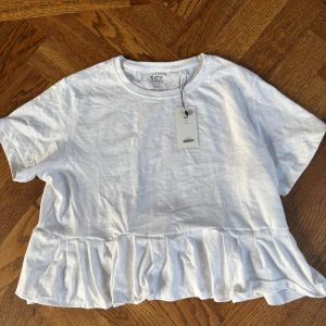 Vit volangtopp från lager 157 - Säljer en söt vit topp från lager 157 med volang nertill. Toppen har rund halsringning och korta ärmar. Perfekt för en fräsch och enkel stil. Den är omsydd från en vanlig T-shirt till en T-shirt med volang där nere. Storlek 160 sitter som xs. Aldrig använd med prislappen kvar