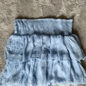 Ljusblå volangkjol med smock - Supersöt ljusblå kjol med smock i midjan och dubbla volanger. Kjolen har ett diskret vitt mönster och är gjord i ett lätt, luftigt material som passar perfekt till varma dagar.