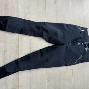 Mörkblå ridbyxor från CRW - Mörkblå ridbyxor från CRW med förstärkta partier på insidan av benen och sätet. Byxorna har två framfickor med dragkedja och bälteshällor i midjan. Perfekta för ridning och har en klassisk, stilren look.