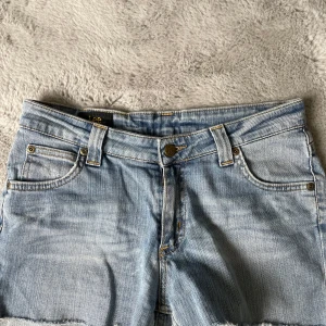 Ljusblå jeansshorts från Lee - Säljer ett par ljusblå jeansshorts från Lee med klassisk femficksdesign och råa kanter nertill. Shortsen har bälteshällor och knappgylf. Perfekta för varma dagar och enkel att matcha med olika toppar.