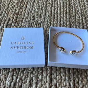Guldfärgat armband med kristaller från Caroline Svedbom - Helt oanvänd! Säljer ett elegant guldfärgat armband från Caroline Svedbom. Armbandet har en tvinnad design och är dekorerat med två stora, klara kristaller i droppformade infattningar. Perfekt accessoar för att lyfta din outfit. Nypris 795kr.