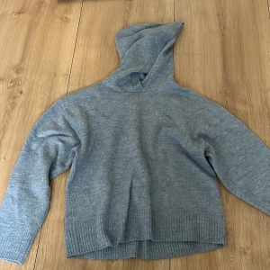 Ljusgrå stickad hoodie från Lindex - Mysig ljusgrå stickad hoodie från Lindex i storlek 146/152. Tröjan har långa ärmar, ribbade muddar och en stor huva. Perfekt för kyliga dagar och enkel att matcha med det mesta.