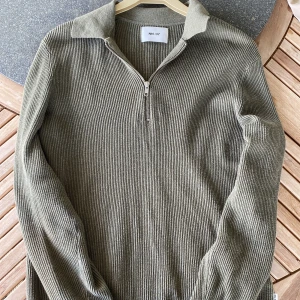 Olivgrön ribbad tröja med dragkedja från NN07 - Snygg olivgrön ribbad tröja från NN07 med halv dragkedja och krage. Tröjan har lång ärm och är tillverkad i ett mjukt material som passar perfekt till alla väder och den en ser sprillans ny ut. Den är lite större i storleken. Nypris 1600kr