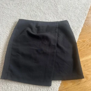 Svart omlottkjol - Säljer en svart kort omlottkjol med stilren design. Kjolen har en rak passform och är perfekt för dig som gillar minimalistiska plagg. Enkel att matcha med olika toppar och skor.