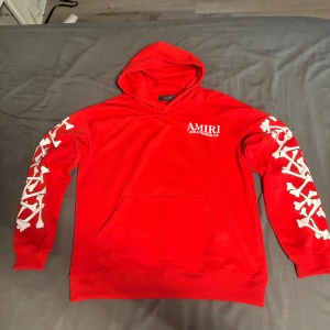 Röd Amiri hoodie - Säljer en röd hoodie från Amiri med vita tryck längs båda ärmarna och logga på bröstet. Tröjan har huva och magficka, perfekt för en streetwear-look.
