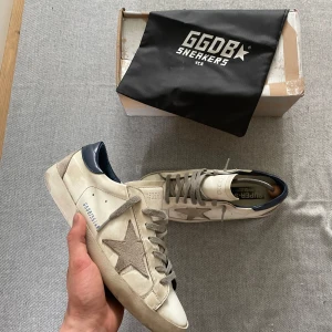 Vita sneakers från Golden Goose - Golden Goose sneakers i vitt skinn med grå snörning och grå stjärndetalj på sidan. Blå hälkappa och platt svart sula. Klassisk modell med rund tå och diskret branding på sidan.