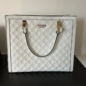 Vit axelväska från Guess  - Säljer en snygg vit axelväska från Guess med präglat logomönster och guldfärgade detaljer. Väskan har svarta handtag, dragkedja upptill och en praktisk ytterficka med dragkedja på baksidan. Perfekt för dig som vill ha en stilren och rymlig väska.