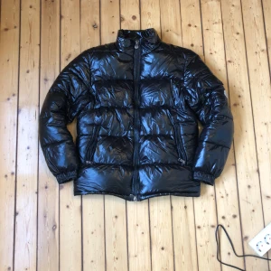 Svart pufferjacka från Moncler - Säljer en svart, glansig pufferjacka från Moncler med hög krage och dragkedja framtill. Jackan har två fickor med dragkedja och en Moncler-logga på ärmen. Perfekt för kyliga dagar. Storlek 1 i moncler storlekar.