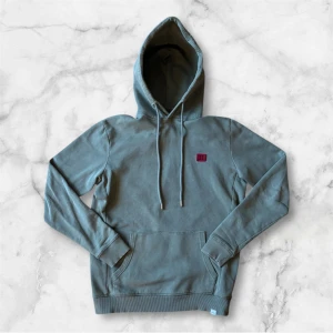 Grön hoodie från Les Deux - Säljer en stilren grön hoodie från Les Deux med röd broderad logga på bröstet. Tröjan har huva med dragsko och en stor magficka framtill. Perfekt för en avslappnad look.