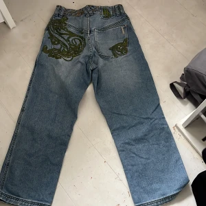 Blå jeans med broderade detaljer från RydelGEAR - Säljer ett par blå jeans från RydelGEAR med unika gröna broderade mönster på bakfickan och sidan. Jeansen har normal passform och raka ben, samt klassiska fickor och bälteshällor. Är 170 och dem är lite korta på mig, skriv om du vill ha fler bilder. Innerbenslängd 69cm. 