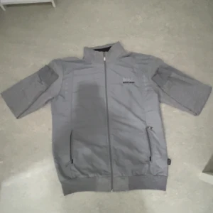 Grå zipjacka från Hugo Boss - Snygg grå zipjacka från Hugo Boss med korta ärmar och broderad logga på bröstet och ärmen. Jackan har dragkedja framtill och två fickor med dragkedja. Perfekt för en stilren och sportig look.