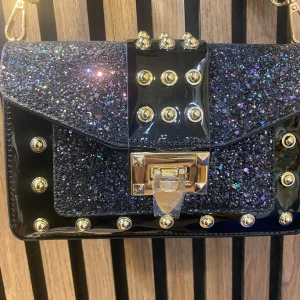 Svart glittrig axelväska med nitar och stjärnmönstrat band - Cool svart axelväska med glittrig yta och guldiga nitar. Väskan har ett brett axelband med stjärnor och text, samt ett snyggt spänne framtill. Perfekt för att lyfta vilken outfit som helst!