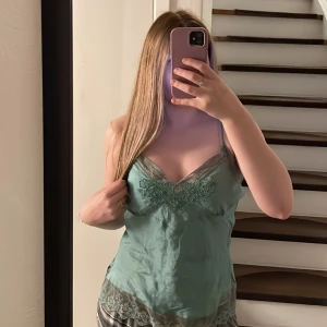 Linne med spetsdetaljer - Säljer en turkos linnetopp med tunna axelband och vackra spetsdetaljer i grått vid halsringning och nederkant. Toppen har en feminin v-ringning och broderade detaljer framtill. Perfekt för sommardagar eller som layering-plagg. Säljer då den ej används