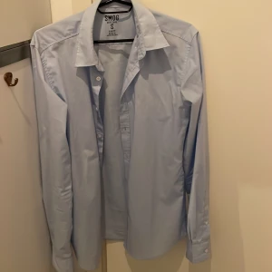Ljusblå skjorta från SMOG - Säljer en ljusblå skjorta från SMOG i slim fit-modell. Skjortan har klassisk krage, långa ärmar och knäppning framtill. Perfekt för dig som gillar en stilren och enkel look.