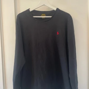 Svart långärmad tröja från Polo Ralph Lauren - Svart långärmad tröja från Polo Ralph Lauren med klassisk röd broderad logga på bröstet. Tröjan har en rund halsringning och är tillverkad i mjuk bomull, perfekt för en stilren och enkel look.