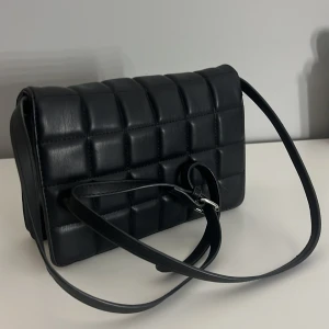 Svart quiltad axelväska - Säljer en svart quiltad axelväska med stilren design och justerbar rem. Väskan har ett rutmönster och är perfekt för att bära dina viktigaste saker. Enkel att matcha med olika outfits.