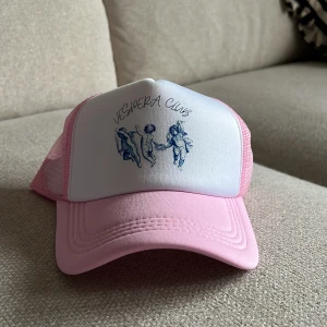Rosa och vit keps med tryck - Snygg keps i rosa och vitt med nät på sidorna och baksidan. Framtill finns ett tryck med blå änglar och texten 'VESPERA CLUB'. Perfekt accessoar för en cool och avslappnad stil.