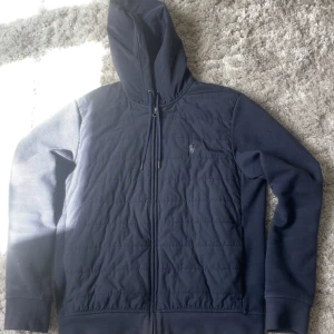 Mörkblå hoodiejacka från Polo Ralph Lauren - Säljer en mörkblå hoodiejacka från Polo Ralph Lauren med dragkedja och quiltad framsida. Jackan har huva med dragsko och broderad logga på bröstet. Ärmar och rygg i mjuk bomullskvalitet, perfekt för lager-på-lager.
