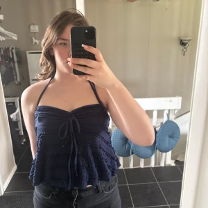 Mörkblå croppad halterneck-top - Säljer en mörkblå croppad halterneck-top med volangdetaljer och knyt framtill. Toppen har en lätt rynkad struktur och tunna band som knyts i nacken. Perfekt för varma dagar och enkel att styla till jeans eller kjol. Passar bra för dig med storlek M/L, det står L på lappen men jag själv har M och den passar bra🩷Inte använd alls förutom nu när jag tog bilderna🫶🏻Kontakta mig för mer info💞