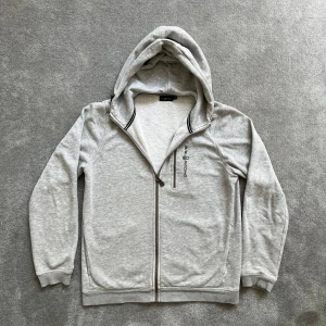 SailRacing Zip Hoodie - Ljusgrå hoodie med dragkedja från Sail Racing! Tröjan har huva med broderad logga samt en mjuk insida. Skick 8/10 då slutet på armarna (muddarna) är lite slitna. Hör gärna av er vid funderingar!