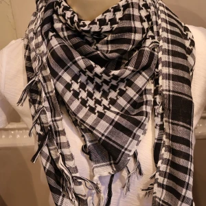 Svartvit rutig halsduk med fransar - Säljer en svartvit halsduk med rutigt mönster och dekorativa fransar längs kanterna. Halsduken har ett klassiskt keffiyeh-inspirerat mönster och är lätt att styla till olika outfits. 100x100cm