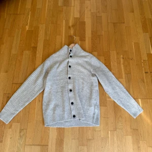 Ljusgrå kofta från Jack & Jones - Ljusgrå ribbstickad kofta från Jack & Jones med hög krage och svarta knappar längs hela framsidan. Tröjan har lång ärm och är tillverkad i ett mjukt material som passar perfekt för kyligare dagar.