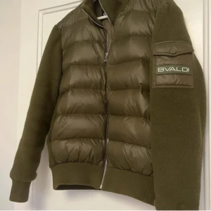 Olivgrön pufferjacka från Bvaldi - Säljer en olivgrön pufferjacka från Bvaldi med stickade ärmar och rygg. Jackan har dragkedja framtill och en ficka med lock och logotyp på ärmen. Perfekt för kyliga dagar och riktigt snygg streetstil.