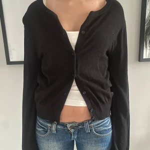 Svart kofta med knappar - Säljer en svart, croppad kofta från HM med rund halsringning och knäppning framtill. Koftan har långa ärmar och är i ett mjukt, tunt material som passar perfekt till lager-på-lager. Enkel och stilren design.