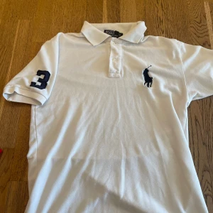 Vit pikétröja från Polo Ralph Lauren - Klassisk vit pikétröja från Polo Ralph Lauren med blå logga på bröstet och siffra 3 på ärmen. Tröjan har krage och knäppning framtill. Perfekt för en stilren och sportig look.