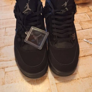 Svarta Air Jordan 4 black cat - Säljer ett par helsvarta Air Jordan 4 black cat (helt nya) sneakers med klassisk siluett och meshdetaljer på sidorna. Skorna har snörning och ikonisk Jumpman-logga på plösen. Perfekta för dig som gillar streetwear och vill ha en stilren look.