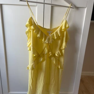 Gul volangklänning med smala axelband - Söt gul klänning med smala axelband och volangdetaljer framtill och längs urringningen. Klänningen har v-ringning och är tillverkad i ett lätt material, perfekt för varma dagar.