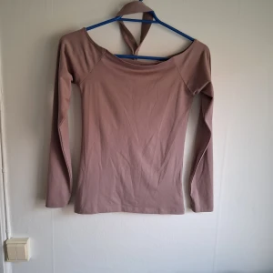 Beige långärmad topp med knytband - Säljer en beige långärmad topp med bred urringning och knytband i halsen. Toppen har en tight passform och är tillverkad i ett mjukt, stretchigt material. Perfekt för att skapa en stilren och trendig look.