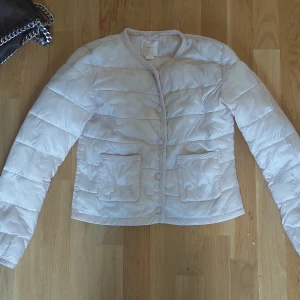Ljusbeige pufferjacka från Gina Tricot - Supersöt ljusbeige pufferjacka från Gina Tricot med quiltad look, två stora fickor framtill och snygga flätade detaljer längs kanterna. Jackan har knappar hela vägen och är perfekt för dig som vill ha en stilren och trendig vibe.