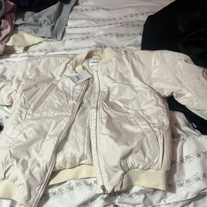 Vit bomberjacka från Kappahl - Snygg vit bomberjacka från Kappahl i storlek 164. Jackan har ribbade muddar, dragkedja framtill och två fickor på sidorna. Tillverkad i glansig polyester och har en clean, stilren look som passar perfekt till streetwear.