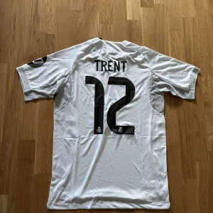 Real Madrid Trent 12 fotbollströja - Snygg vit Real Madrid fotbollströja från Adidas med svarta detaljer och guldiga linjer längs sidorna. Tryck med 'Trent' och nummer 12 på ryggen, samt klubbmärke och sponsor på framsidan. Tillverkad i lätt och ventilerande polyester, perfekt för match eller träning.