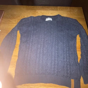 Mörkblå stickad tröja från 157 - Mörkblå kabelstickad tröja från 157 med rund hals och ribbade muddar. Tröjan har ett snyggt mönster och en liten läderdetalj nertill. Perfekt för kyliga dagar och passar till det mesta i garderoben.