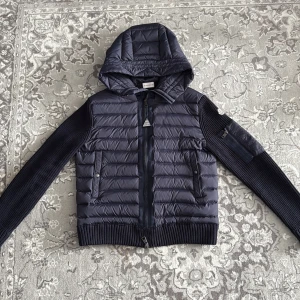 Mörkblå Moncler pufferjacka med huva - Snygg mörkblå pufferjacka från Moncler med stickade ärmar och quiltad kropp. Jackan har huva, dragkedja framtill och två fickor med knappar. Moncler-logga på ärmen och ribbade muddar ger en clean och exklusiv look.