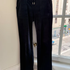 Mörkblå velourbyxor Juicy Couture XS - Mörkblå mjukisbyxor från Juicy Couture i glansig velour. Byxorna har raka ben, snörning i midjan och stora fickor både fram och bak med tryckknappar. Materialet är 95% polyester och 5% elastan för extra stretch och komfort.